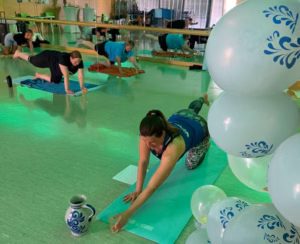Yogakurs in einem hellen Kursraum mit Spiegelwand: Im Vordergrund streckt sich eine Frau auf einer grünen Matte nach vorn und greift nach einem Glas, daneben steht ein blau-grauer Bembel. Im Hintergrund machen weitere Teilnehmende Übungen im Vierfüßlerstand. Rechts stehen weiße Luftballons mit blauem Bembel-Muster.